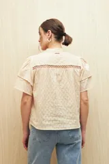 Camisa de algodón color beige con cuello mao, cierre frontal de botones y mangas cortas con volados. Presenta un diseño texturizado con rayas verticales y detalles de pasamanería calada en el frente.