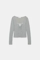 Chaqueta de punto color beige con escote en pico y manga larga, que incluye un top interior integrado con detalle de encaje a tono. Cierre frontal mediante botones.