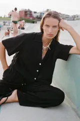 Pantalón negro de tiro alto con cintura elástica ajustable mediante cordones con terminales dorados. Presenta pernera ancha y bolsillos laterales. La tela tiene una textura sutilmente arrugada o plisada.