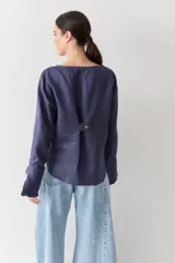 Blusa de lino con cuello bote y mangas largas terminadas en puños acampanados con volados. Presenta un cierre con botón en la espalda.