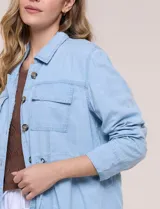 Sobrecamisa azul de denim de algodón liviano, con cuello camisero, cierre frontal de botones, bolsillos con solapa en el pecho y cordón ajustable en la cintura.