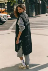 Chaleco largo acolchado de color negro, con cierre frontal completo y diseño sin mangas. Presenta un acabado matelasse y un borde contrastante en color claro a lo largo de la cremallera.