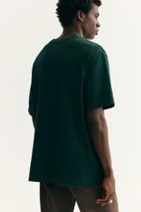 Remera verde oscuro con estampado frontal de Snoopy, equipaje y la inscripción "More Places - Take (more) care with Peanuts". Cuenta con cuello redondo y mangas cortas con hombros caídos.