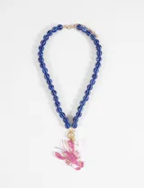 Collar de cuentas redondas de vidrio azul oscuro, con dije colgante de langosta en color rosa fucsia y un dije de perla blanca. El cierre es de metal dorado.