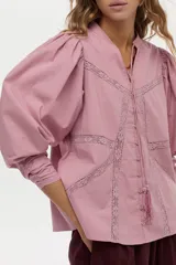 Blusa de algodón liviano color blanco, con detalles de puntilla en el frente, cuello mao con escote en V, cordón colgante y mangas largas abullonadas con puños ajustados.