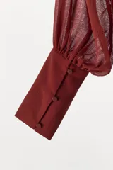 Vestido largo fluido en color bordó, confeccionado en georgette. Presenta cuello redondo, mangas largas con pliegues terminadas en puños y tiras ajustables para atar en la cintura. Incluye forro interior a tono.