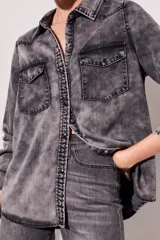 Camisa de denim celeste con cuello clásico, cierre frontal con botones a presión, dos bolsillos con solapa abotonada en el pecho y mangas largas con puños abotonados.