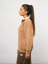 Buzo de felpa color tostado con cuello alto y medio cierre frontal. Presenta un diseño de corte holgado con hombros caídos, puños y ruedo acanalados, y el logo de la marca bordado en el pecho.