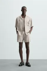 Camisa de lino de calce relajado en color beige, con cuello bowling y manga corta. Cuenta con dos bolsillos de parche en el pecho, aberturas laterales en el bajo y cierre frontal con botones.