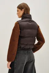 Chaleco acolchado de corte corto, color verde oliva, con cuello alto ajustable mediante cordones elásticos y cierre frontal con cremallera.