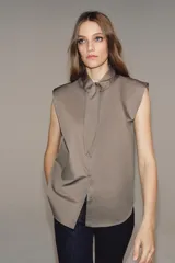 Camisa sin mangas confeccionada en algodón, con cuello redondo y detalle de lazada frontal. Presenta cierre frontal con botones ocultos por solapa y bajo redondeado.