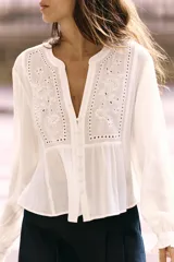 Blusa blanca de manga larga con escote en pico y cuello redondo. Cuenta con bordados florales frontales a tono, puños con volados y cierre frontal de botones.