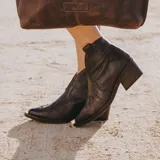 Botín de cuero vacuno color marrón oscuro, con taco de madera foliado en cuero de 5cm de altura, cierre lateral interno y horma de punta fina.