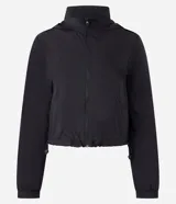 Campera cortavientos deportiva de corte cropped, color verde oscuro. Presenta capucha, cierre frontal con cremallera, mangas largas con puños elásticos y cordón ajustable en el ruedo.