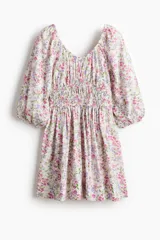 Vestido corto de algodón con estampado floreado en tonos rosados y lilas sobre base blanca. Presenta escote en V fruncido, mangas globo a los codos con puños elásticos y sección fruncida en la cintura.
