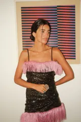 Vestido corto sin mangas, color negro, completamente cubierto de lentejuelas brillantes. Presenta un borde de flecos largos de color rosado en el escote y en el ruedo inferior.