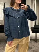 Camisa de manga larga en color azul oscuro con textura cuadriculada. Presenta volados en la parte superior delantera y en los puños, cierre frontal con botones y cuello redondo.