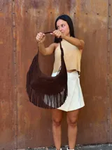 Cartera de hombro tipo hobo confeccionada en cuero gamuzado color chocolate, con detalle de flecos largos en la parte inferior y correa ajustable con hebilla metálica.