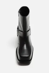 Bota corta negra de cuero con horma cuadrada, tacón bajo y arnés decorativo con remaches plateados a la altura del tobillo.