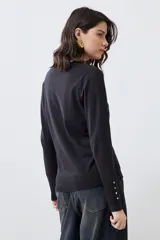 Cardigan de punto negro con escote en V, cierre frontal con botones metálicos dorados y mangas largas con puños ajustados que incluyen detalles de botones decorativos.
