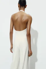 Mono largo de color blanco con cuello halter y espalda descubierta. Presenta un diseño de pierna ancha y cierre posterior con cremallera oculta y botones.