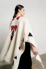 Ruana tejida en color crudo con diseño geométrico inspirado en motivos Navajo en tonos bordó y negro. Presenta terminaciones con flecos en el ruedo y un pequeño aplique de cuero en la parte inferior.