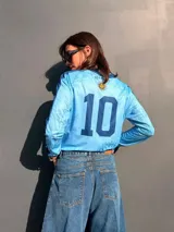 Remera de manga larga en color celeste con estampado de soles y flores de ceibo en tono azul. Presenta cuello polo y puños en tejido rib azul marino, con un parche circular bordado en el pecho que incluye el sol y la inscripción Uruguay Vdamiani.