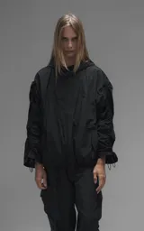 Campera rompevientos negra de calce oversize, confeccionada en microfibra técnica ligera con acabado semi-mate. Presenta capucha integrada, cierre frontal técnico y bolsillos tipo ojal. Destaca por su sistema de frunces elásticos regulables mediante tancas a lo largo de las mangas y el ruedo, permitiendo ajustar el volumen de la prenda.