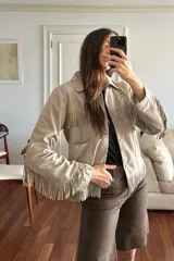 Campera de gamuza color crudo con flecos en el pecho, en el ruedo y en las mangas, cuello camisero, bolsillos frontales appliqués y cierre con broches a presión oculta.