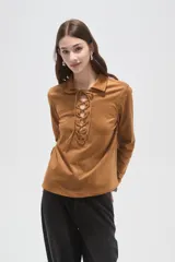 Blusa de gamuza sintética color camel, con cuello camisero y cierre frontal mediante cordones entrelazados. Presenta mangas largas y un corte holgado.
