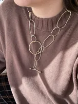 Set de accesorios compuesto por un sobre organizador de gamuza color marrón, un collar de cadena dorada, dos pares de caravanas colgantes y un prendedor, todos en metal dorado.