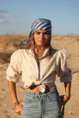 Camisa de lino color beige con mangas globo y detalles de vivos estampados a rayas en azul y marrón. Presenta cierre frontal con botones y diseño anudable en la cintura.