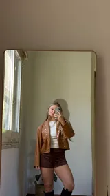 Campera corta de ecocuero color camel, con diseño de cuello solapa y cierre frontal con botones metálicos a presión. Presenta detalles de tachas metálicas a lo largo de la solapa y en el ruedo.