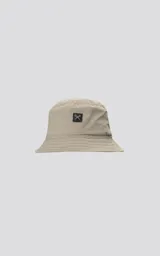 Gorro tipo bucket beige, con ala ancha y costuras horizontales. Presenta un pequeño parche cuadrado negro con un logo de tijeras cruzadas en blanco en el frente.