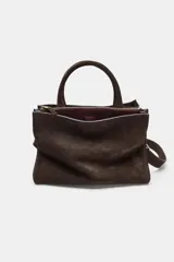 Bolso de mano formato city confeccionado en gamuza color marrón oscuro. Cuenta con doble asa de mano, asa bandolera extraíble, compartimento interior con cierre y bolsillo exterior con imán.