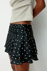 Pollera mini de satén negro con estampado de lunares blancos. Presenta una cintura ancha con pliegues horizontales y un diseño escalonado con volados que aportan volumen y movimiento.