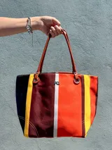 Cartera tote Cano confeccionada en canvas con patrón de rayas verticales multicolor en tonos rojo, marrón, amarillo, naranja y negro. Cuenta con asas de hombro de cuero sintético marrón, detalles de argollas metálicas y logo de la marca metálico al frente.