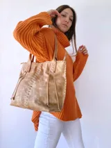 Cartera tipo tote de cuero color camel, con diseño artesanal que presenta costuras decorativas visibles en los laterales y el frente. Cuenta con doble asa de hombro y cierre superior.