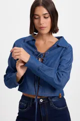 Camisa crop de denim azul con cuello camisero y cierre frontal mediante cordones ajustables entrelazados en ojales. Presenta mangas largas con puños rectos y corte entallado.