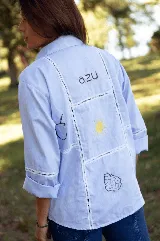 Camisa de lino color celeste, con cuello clásico, cierre frontal de botones y detalle de cuatro estrellas bordadas en color amarillo sobre el pecho. Presenta mangas tres cuartos con detalle de encaje en los puños.
