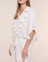 Blusa blanca de silueta holgada con sutil trama en hilo metalizado lurex, escote en V con pechera tejida en crochet y apliques de borlas en el contorno del cuello y las mangas.