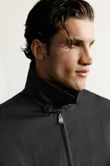Cazadora negra de corte regular fit, confeccionada en tejido técnico con mezcla de algodón. Cuenta con cuello solapa, manga larga con puño abotonado, bolsillos de vivo en la cadera, pliegues laterales en la espalda y bajo elástico. Cierre frontal con cremallera.