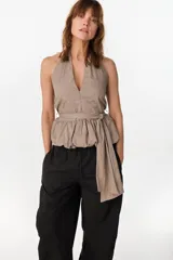 Blusa de cuello halter en color beige, con escote en V pronunciado, corte ajustado en el torso y detalle de volados fruncidos en la cintura con un lazo lateral ajustable.