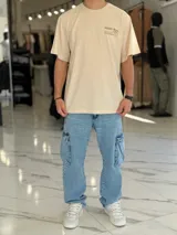 Remera oversize color beige, con mangas cortas y cuello redondo. Presenta un diseño estampado con texto en la parte trasera y lateral, de corte holgado y estilo urbano.