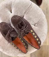 Náuticos Antonia de gamuza color marrón, con flecos decorativos en la parte superior, cordones frontales y detalle de tachas metálicas doradas a lo largo de los laterales.