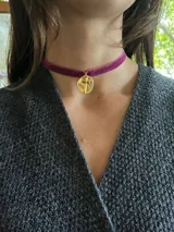 Gargantilla tipo choker de cinta de terciopelo color fucsia con dije circular metálico dorado que presenta una cruz en relieve.