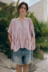 Top blanco de cuello redondo y mangas tipo capa, con textura fruncida, bajo asimétrico, lazos decorativos en el escote y en los laterales, y cierre posterior con botón.