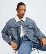Campera trucker de jean azul con cuello de corderito blanco, cierre frontal con botones metálicos, bolsillos frontales con solapa y puños abotonados.