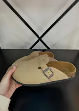 Zuecos de gamuza color beige con hebilla metálica lateral ajustable y suela de corcho con base de goma negra.