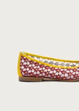 Bailarina de diseño artesanal confeccionada en rejilla bordada con patrón floral en tonos rojizos sobre fondo claro. Presenta ribetes y talón en color amarillo vibrante, con un detalle de media esfera metálica dorada en la puntera y suela plana.